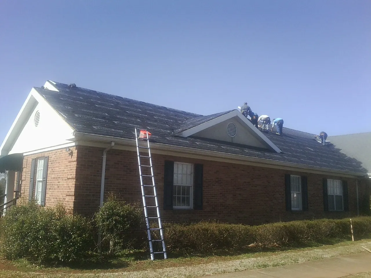 Crew tearing shingles off a brick ranch roof with a ladder set — Metal Roof Repair work by our crew in Left Hand