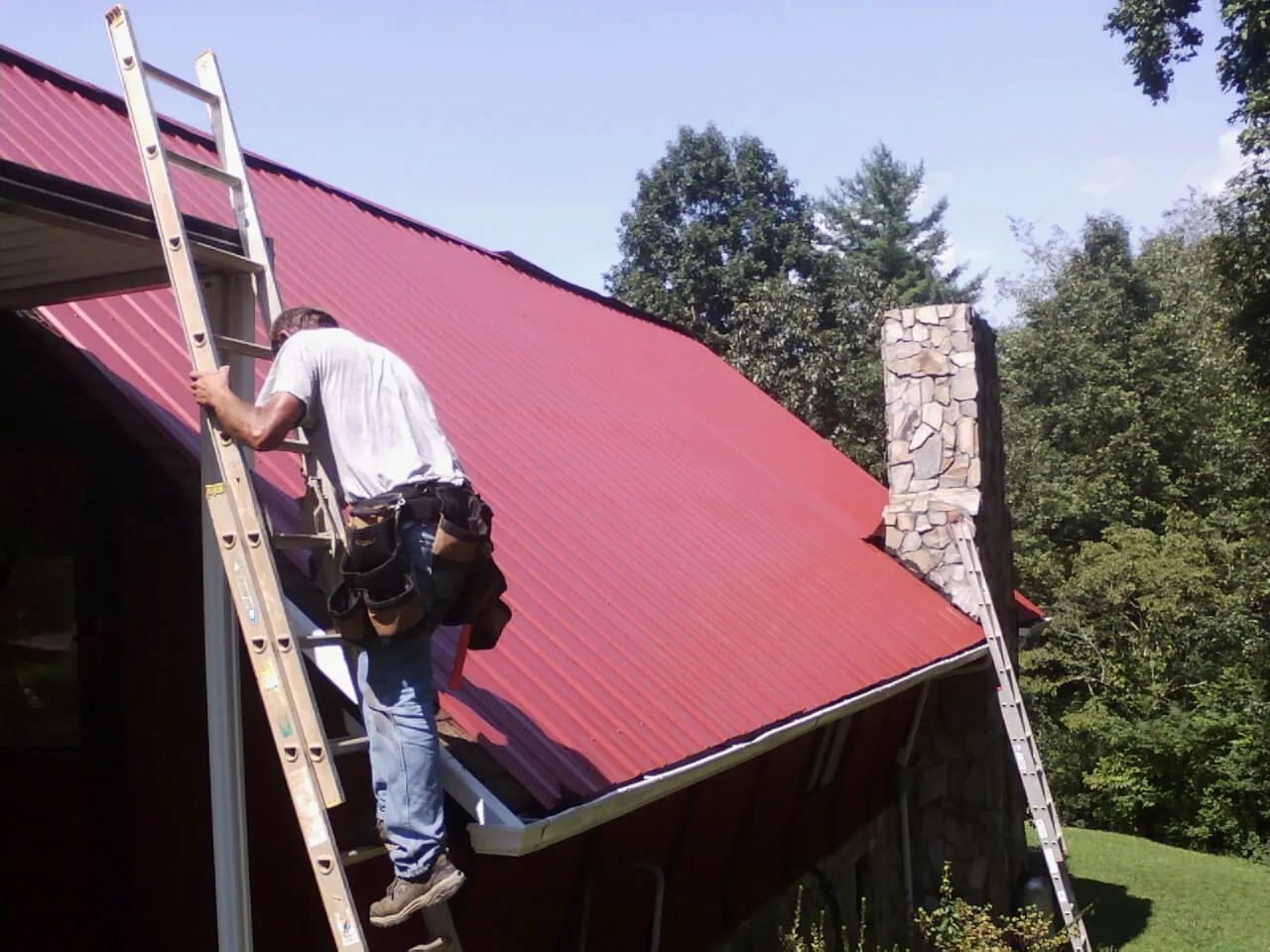 Roofer on a ladder beside a red metal roof and stone chimney — Soffit & Fascia Repair by Roofing Brothers in Left Hand